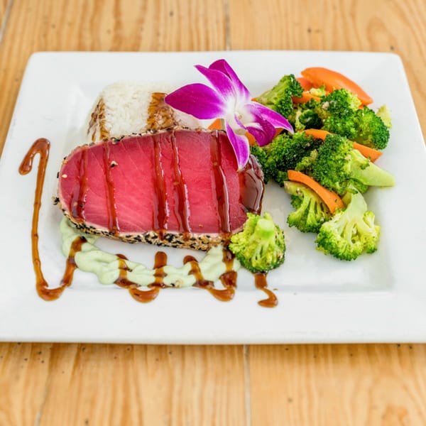 Seared tuna with sesame crust, rice, broccoli, carrots, and a purple orchid on a white plate.