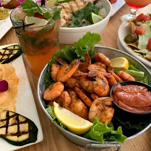 A bucket of grilled shrimp with cocktail sauce, lemon, and lettuce, alongside drinks and other dishes.