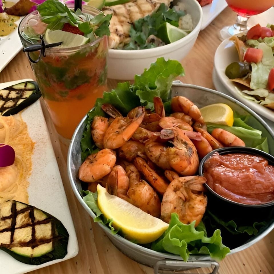 A bucket of grilled shrimp with cocktail sauce, lemon, and lettuce, alongside drinks and other dishes.