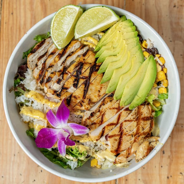 Grilled chicken, sliced avocado, lime wedges, and rice with a purple orchid in a white bowl.